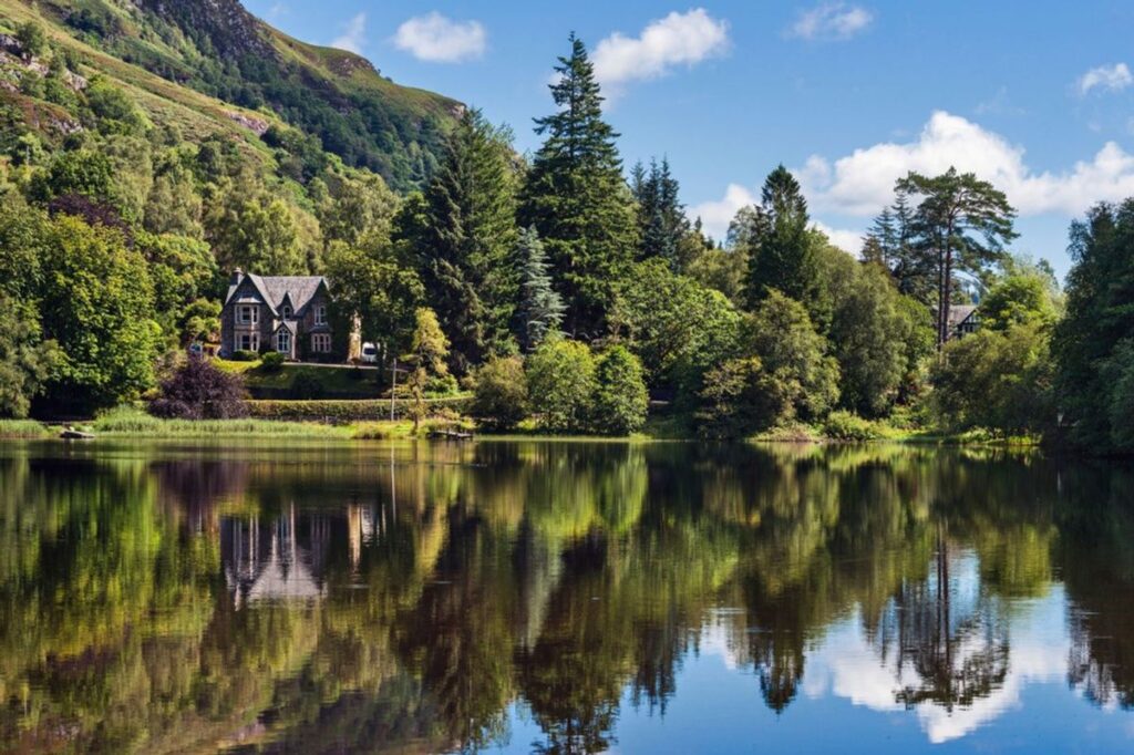 A serene lakeside view showcasing a reflection of trees and a solitary Scottish farmhouse amidst lush greenery.