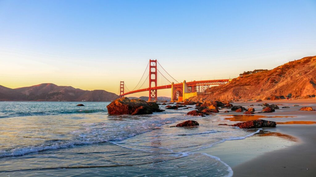 Golden Gate Bridge at sunset viewed from a beach with gentle waves and rocks in the foreground while travelling with your sibling.