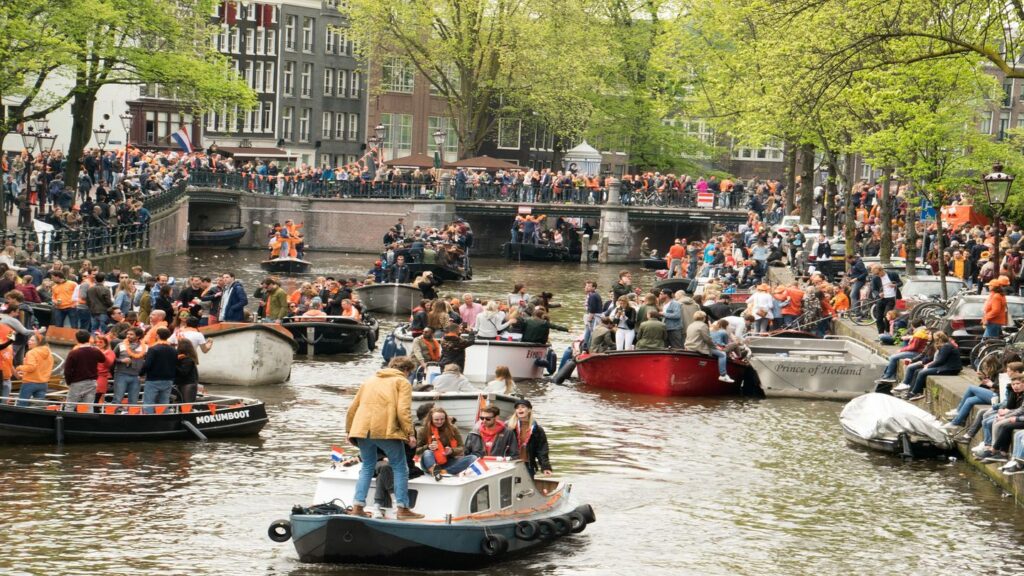 Crowds of people in boats and surrounding the canal celebrate King's Day in Amsterdam. Many are wearing orange in celebration.