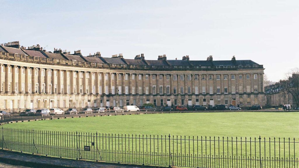 Sweeping crescent of historic Georgian townhouses, reminiscent of the Bridgerton series 2 setting, with parked cars in front, behind a green lawn and metal fence.