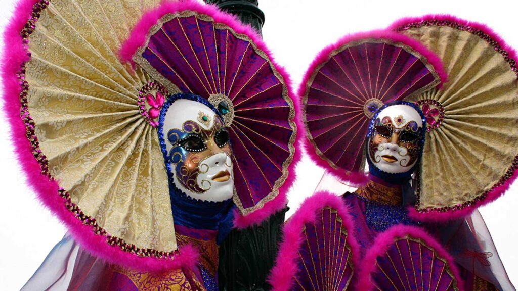Two people in ornate Venetian masks and costumes with large, fan-like collars in purple and gold, embody a scene straight from the bucket list events of Carnival.