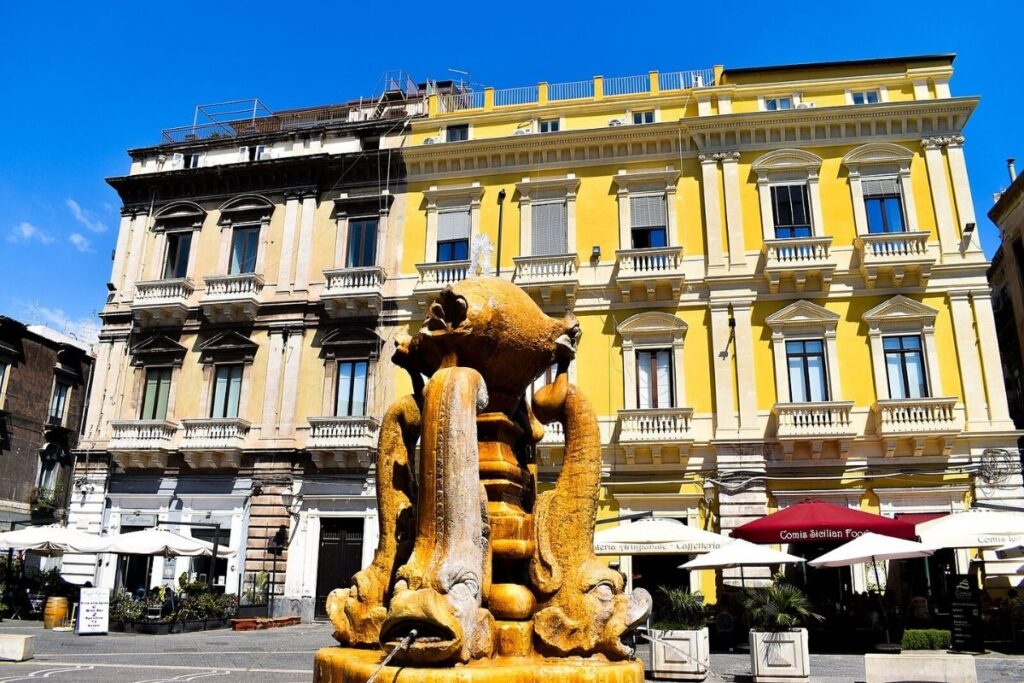 Front of the Fontana dei Delfini and building on Piazza Vincenzo Bellini in Catania, fountain with fish sculptures in front of three buildings, two yellow and one neutral coloured. Umbrellas and dining area in front of the building.