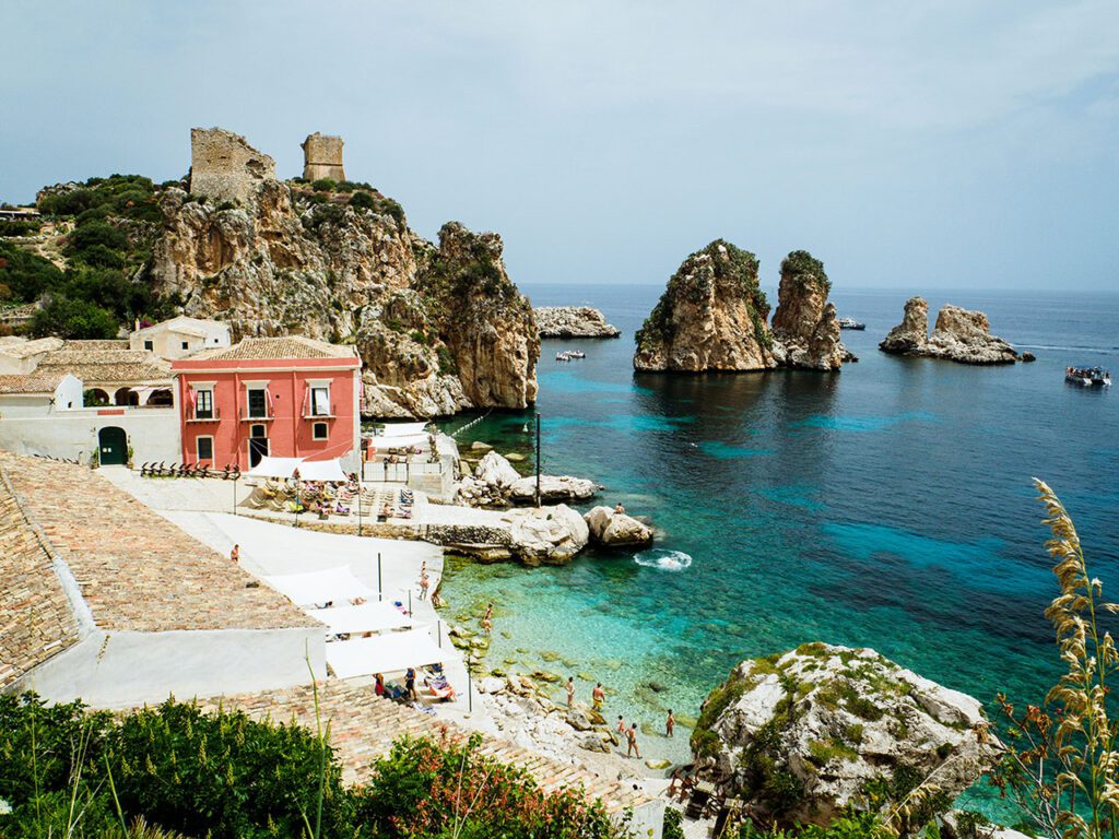 Scopello scenery, buildings on the left hand side with a white rocky sand beach below, with blue clear waters on the right hand-side.