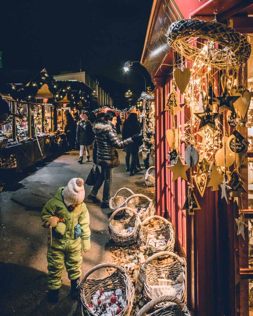 A child in a winter suit explores a festive market stall adorned with lights and Christmas decorations at one of the Christmas Markets of Europe at night.