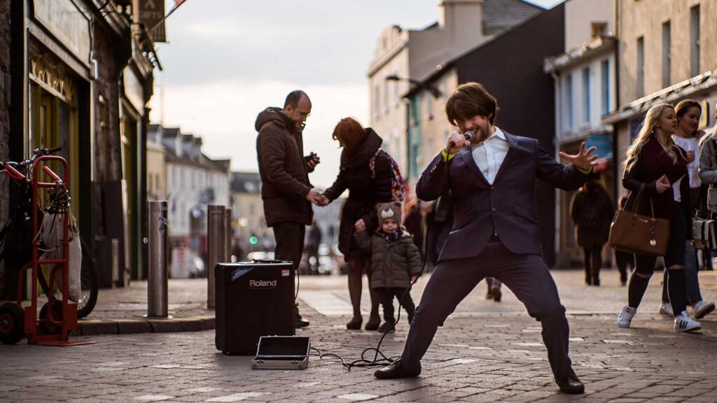 A busker, holding a microphone, strikes a pose in a town street with people walking by 