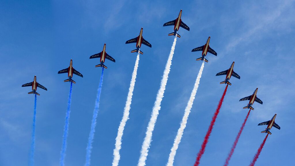 Eight jets flying in formation against a clear blue sky to celebrate Bastille Day, trailing multicolored smoke in blue, white, and red.