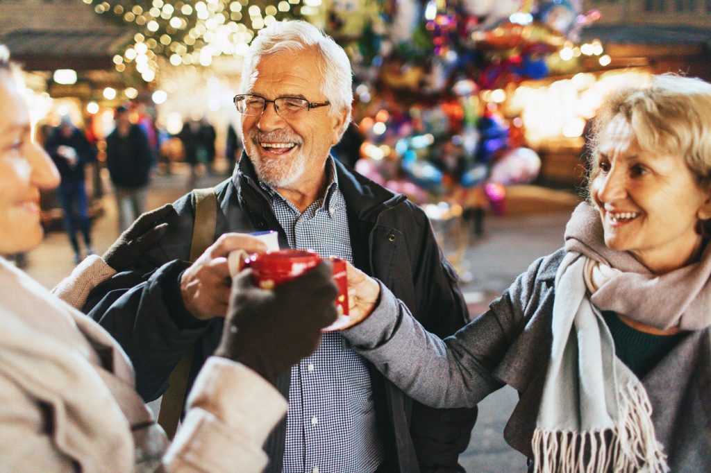 Older couple enjoying mulled wine with a friend at a bustling night market in one of the best places to retire abroad, smiling warmly under festive lights.