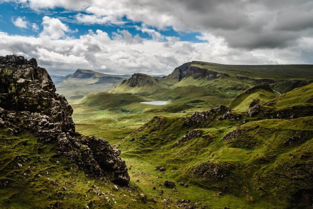 Vast green landscape featuring rugged terrain with rocky outcrops, patches of grass, and a small lake under a cloudy sky, ideal for tracing your heritage.