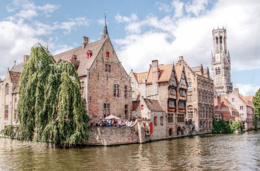 Historic brick buildings and a tall, slender tower beside a tranquil canal, populated with lush trees and a clear sky, in Bruges, Belgium, perfect for your winter travel bucket list.