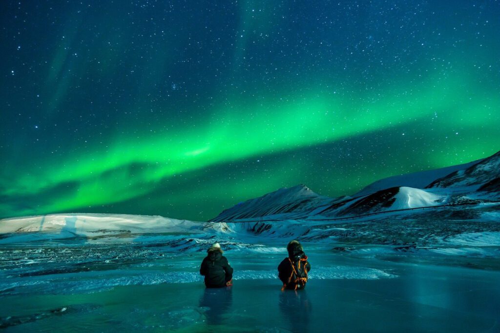 Two people sitting on a frozen lake watching the natural wonders of the northern lights over snow-covered mountains at night.