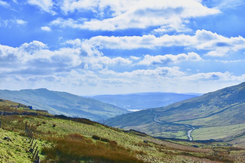 Sunny landscape showing a green valley, winding road, and mountains under a cloudy blue sky during the autumn equinox in England's Lake District.