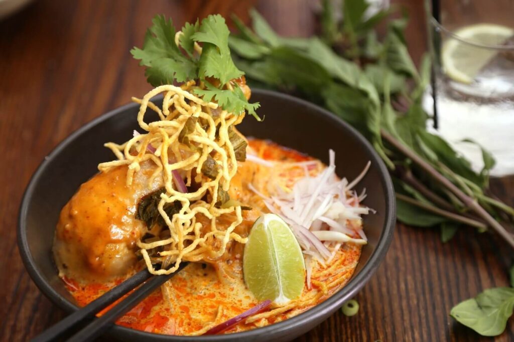 A bowl of khao soi with chicken, topped with crispy noodles and cilantro, served with lime and side herbs on a wooden table, is considered one of the spiciest dishes.