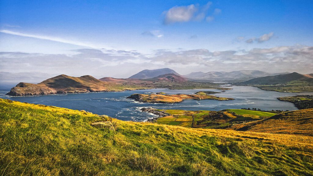 Lush green landscape with rolling hills beside a calm river, leading to distant mountains under a clear sky, perfect for 2023 tours.