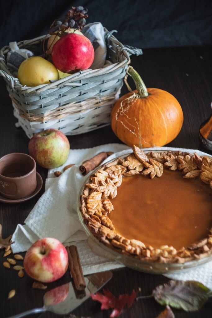 A freshly baked pumpkin pie with decorative crust beside a basket of assorted fruits and a whole pumpkin on a dark wooden table, prepared for Canadian Thanksgiving.