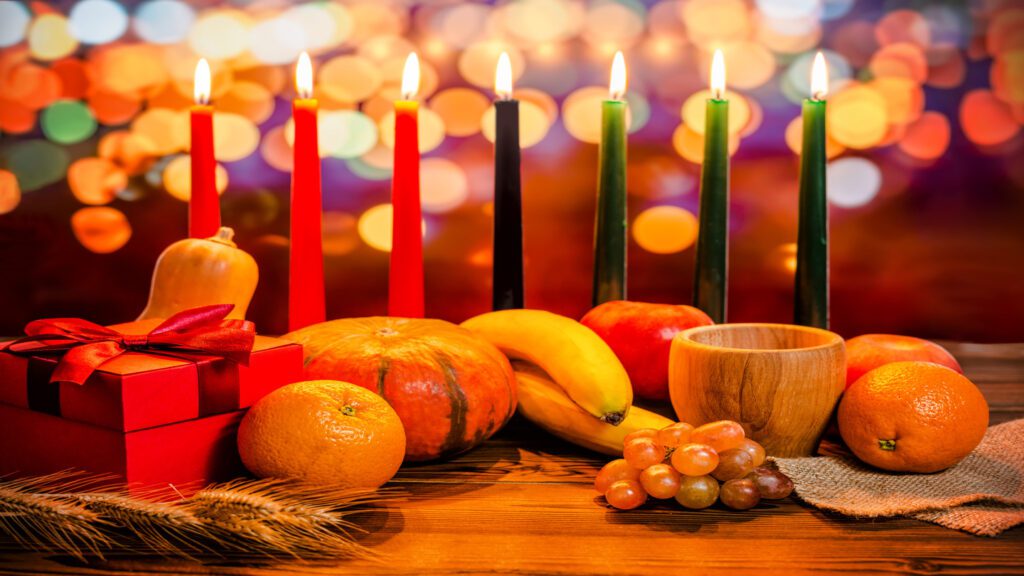 kinara vegetables fruit gifts Kwanzaa. The seven candles of Kwanzaa are in the background, behind fresh fruit laid on a table. The first three candles on the left are red, the middle is black, and the three on the right are green.