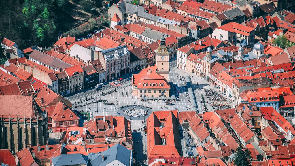 Aerial view of a Balkan town square with red-roofed buildings and a crowd gathered around a central monument.