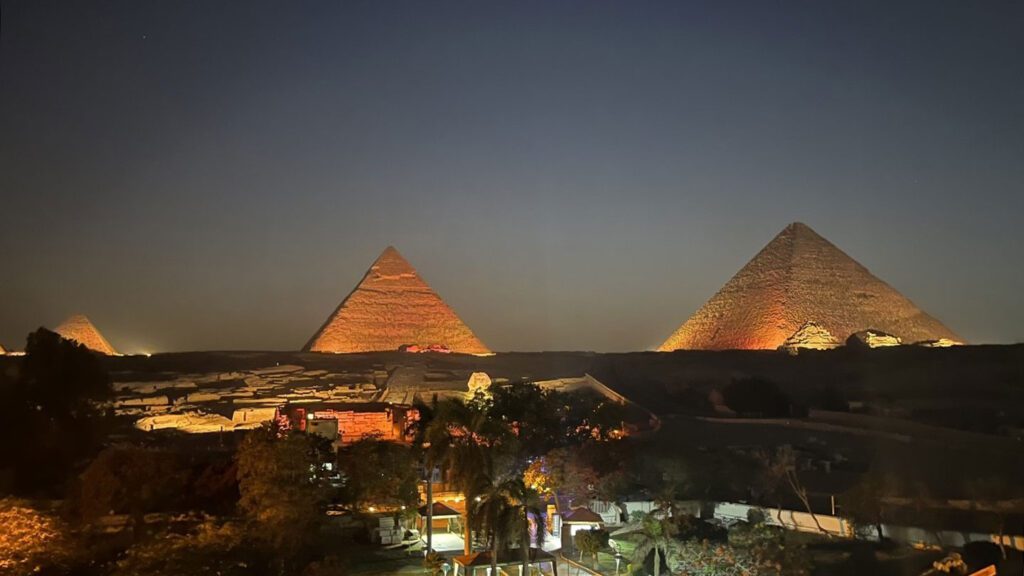 Nighttime view of the great pyramids of Giza, Egypt, illuminated, with a clear sky and surrounding landscape visible.