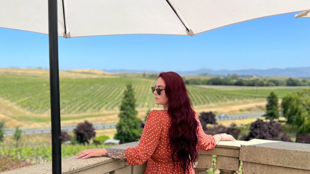 A woman with red hair wearing sunglasses and a polka dot dress looks out over a vineyard from under a patio umbrella on her romantic European tour.