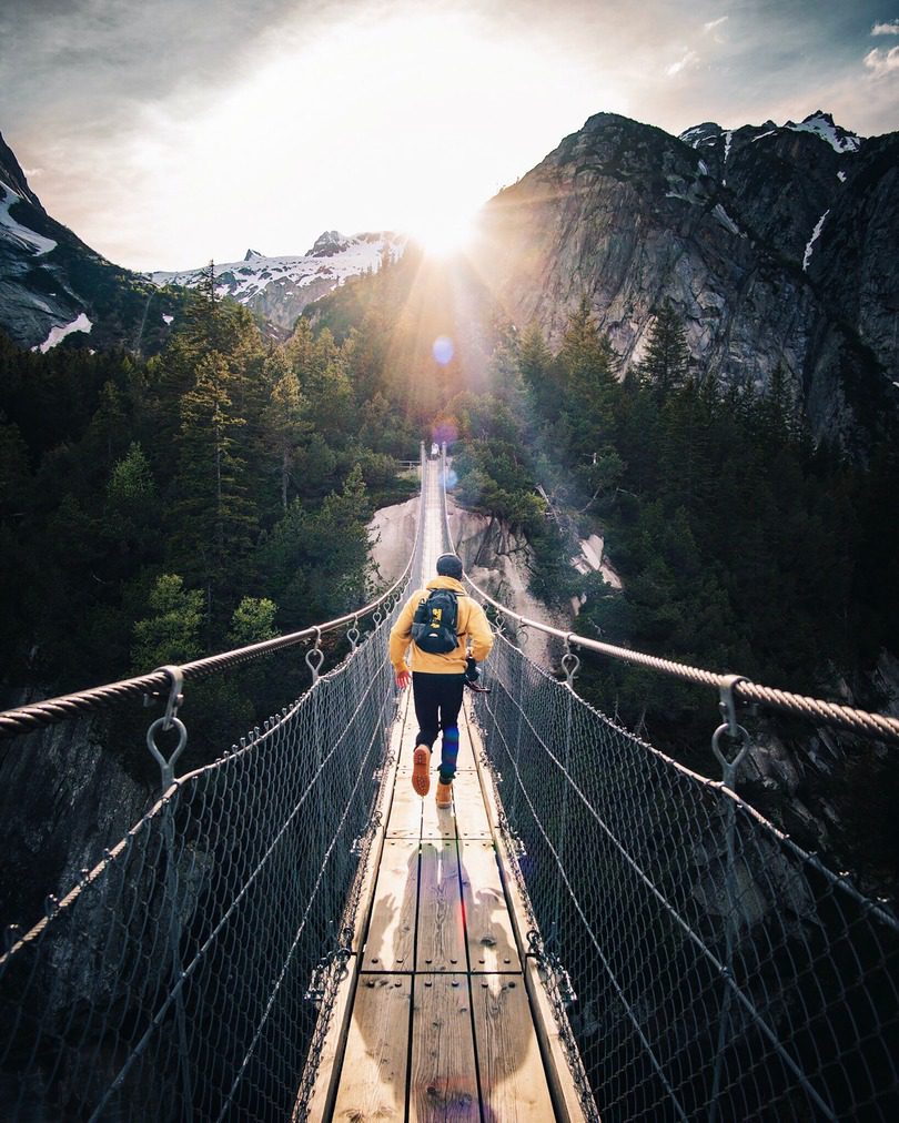 traveller walking over a wooden bridge surrounded by rocky mountains and green forest