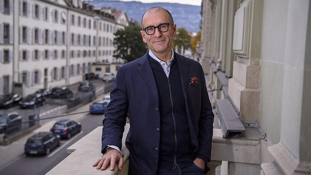 Portrait of Gavin Tollman. He's standing on a balcony overlooking a street in Geneva. He's wearing dark-framed glasses and a blue jacket over a navy sweater with a white shirt underneath.