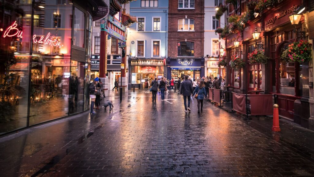 A bustling evening scene on a wet cobblestone street lined with London's top theaters and restaurants, with people walking and a bird in flight.
