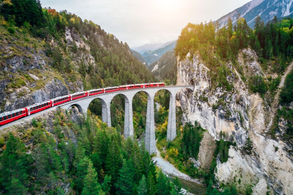 Welcome to Switzerland: A red train crossing an arched stone bridge over a gorge, surrounded by forested mountains during sunset.