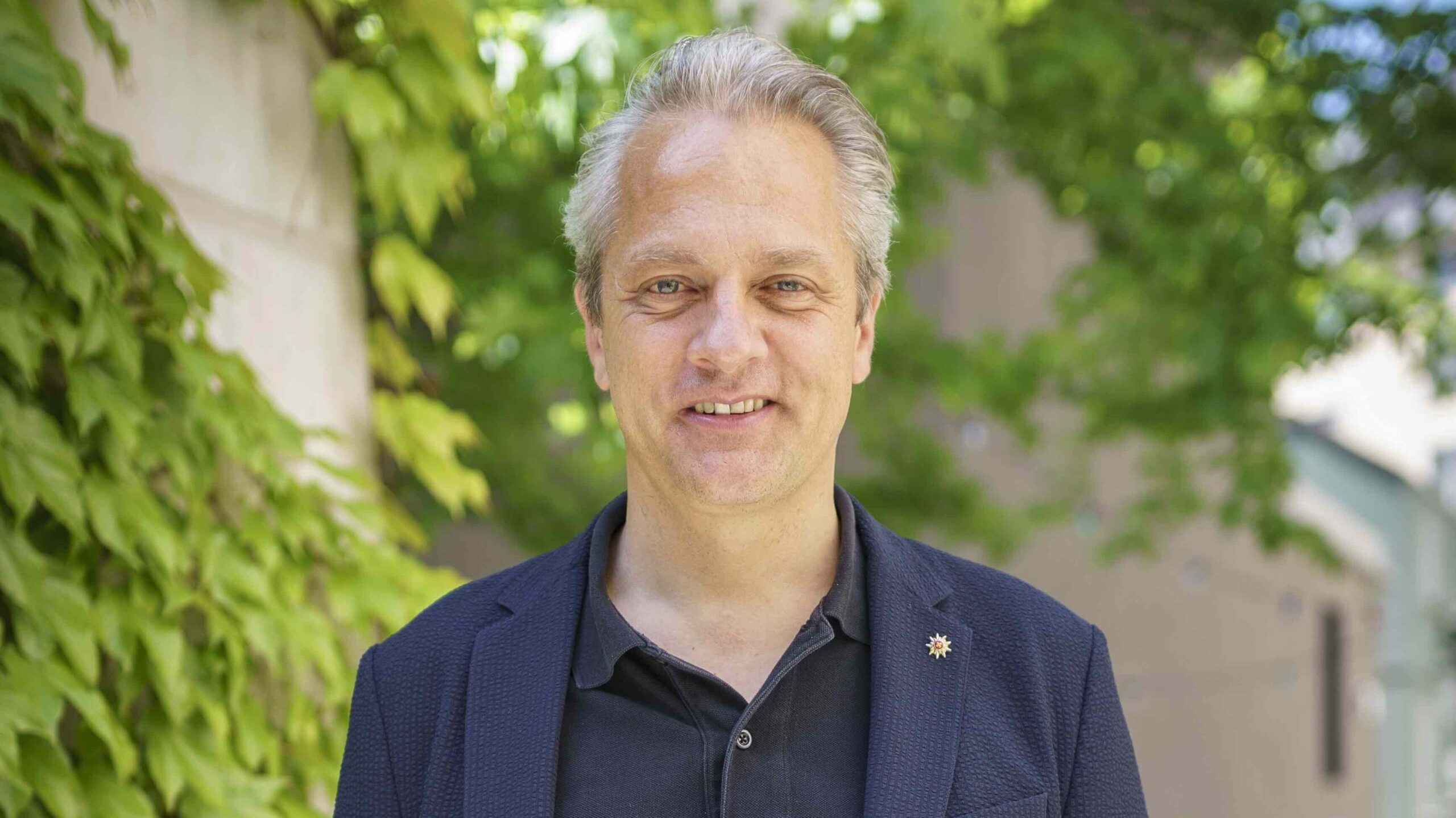 Portrait image of Simon Bosshart. He's standing outside, against a background of a leafy street. He's wearing a blue blazer over a black shirt.