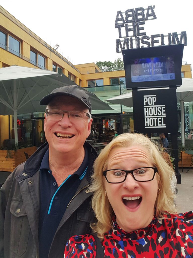 Leith and David selfie image in front of the Abba Museum sign. David is wearing a black cap and glasses; Leith has blonde hair and glasses