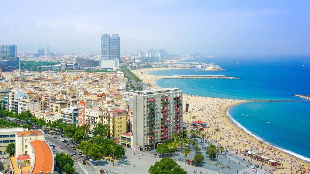 Aerial view of a crowded beach alongside a cityscape with modern buildings and a clear blue sea under a hazy sky, perfect for city travel.