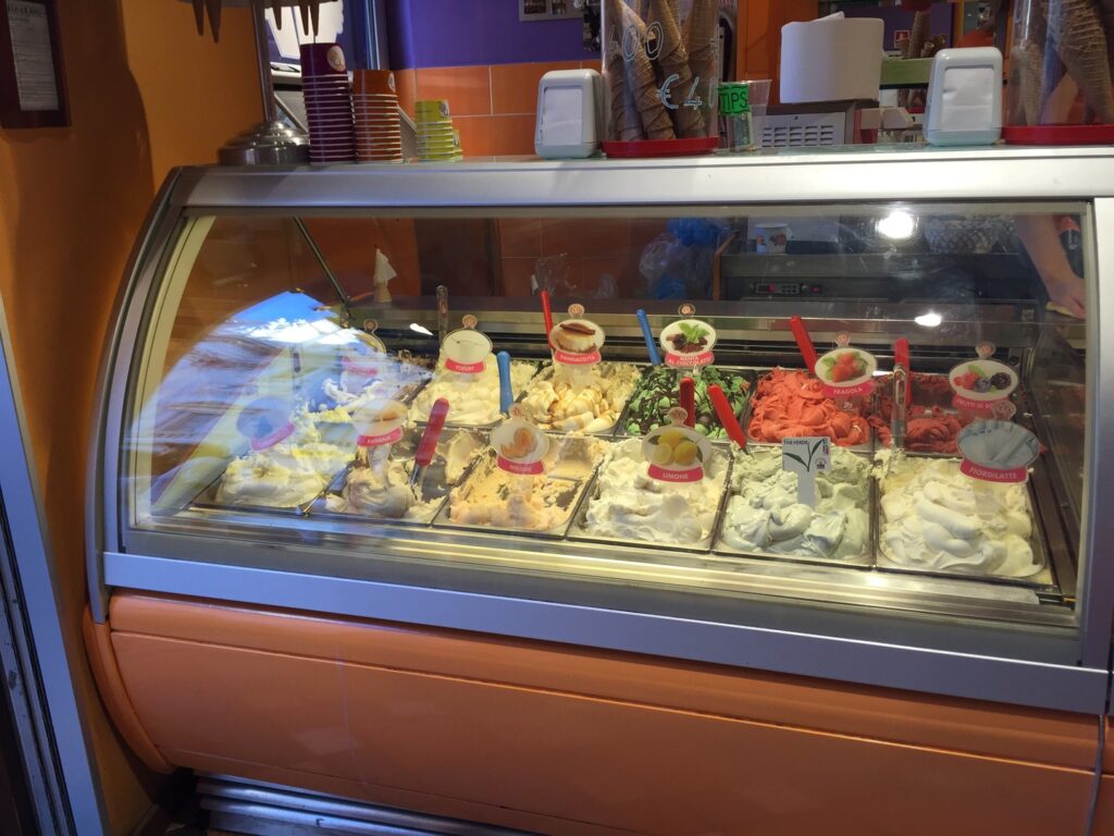 A gelato shop display case in Venice featuring a variety of colorful gelato flavors, each topped with a descriptive label and some garnished with fruit.