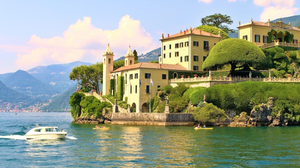 View of Lake Como and one of its opulent villas, with a speedboat in the foreground and mountains in the distance. 