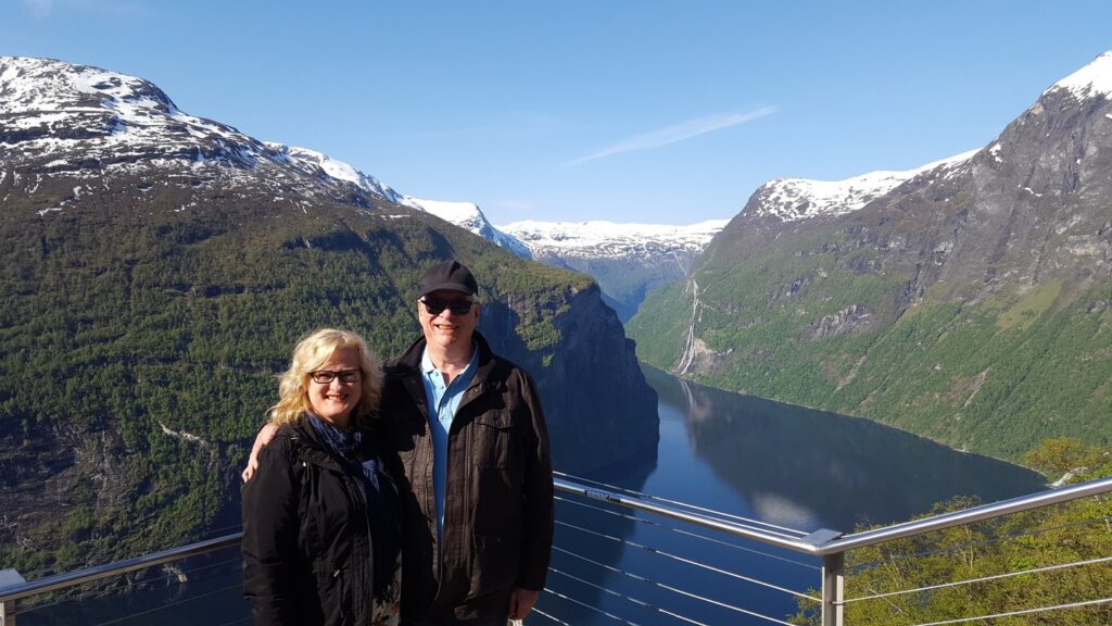 Image of Leith and David standing in front of one of Norway's fjords; the tops of the peaks on both sides of the fjord are covered in snow; sun is illuminating their faces
