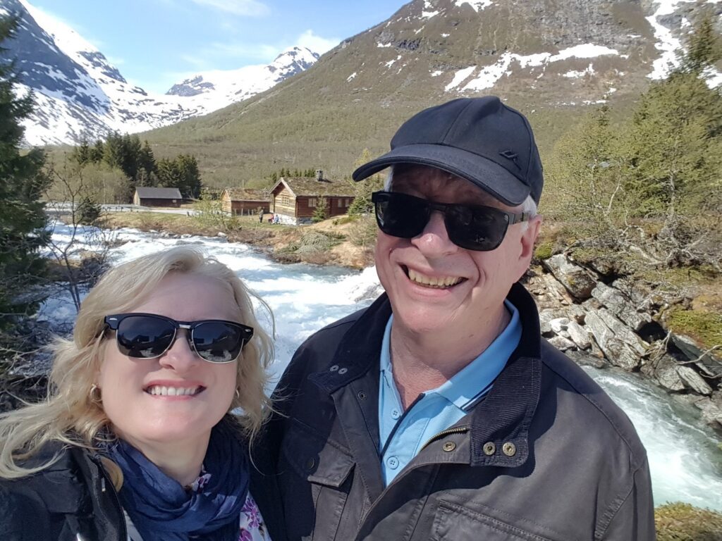 Selfie image of Leith and David standing in front of a river in a mountainous landscape, with log cabins in the background. David is wearing a black cap and sunglasses; Leith is also wearing sunglasses