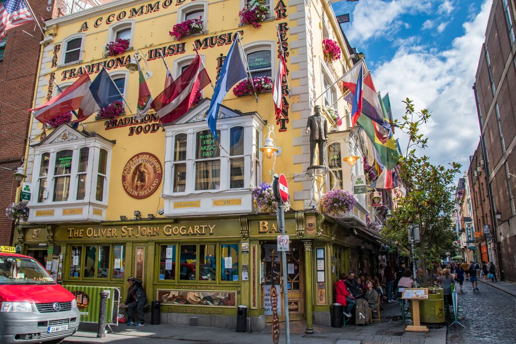 Yellow building with multiple flags, labeled "the oliver st. john gogarty," in an urban area with pedestrians and parked cars, prominently featuring a Trafalgar Tours sign.