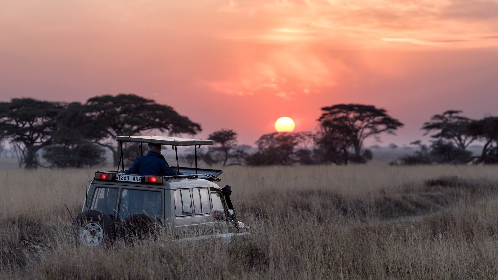 Photo of safari jeep parked in long grass looking towards the sun setting behind some trees