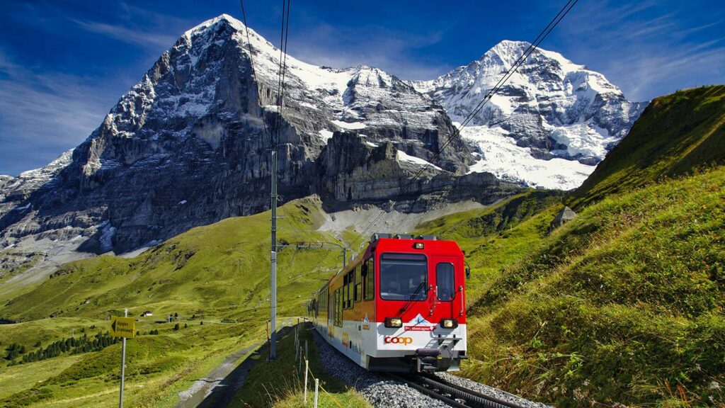 Image of a red mountain train winding through green hills with a snow-capped mountain in the background