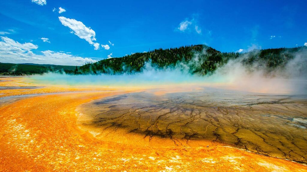 Image of Yellowstone National Park's Grand Prismatic Spring. Steam rising from bright orange water, against a blue sky with green forest-covered hills in the background