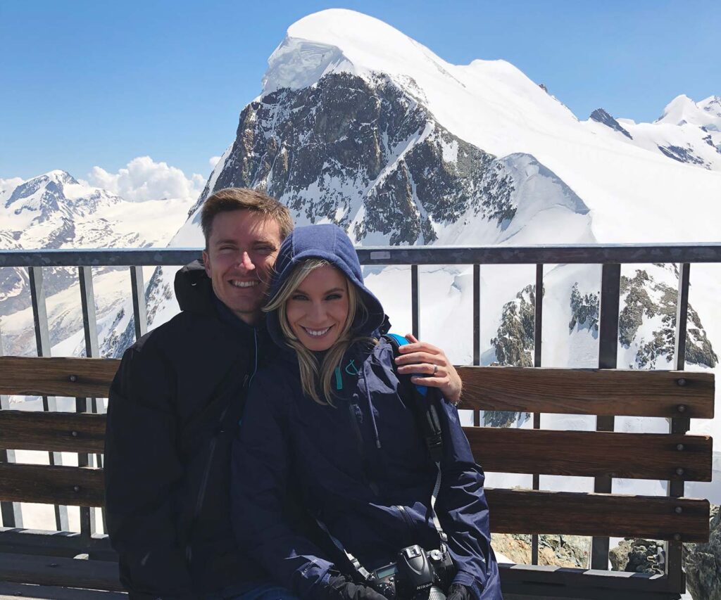 Ashlinn and her husband sit side by side on a bench against a backdrop of snow-covered mountain peaks