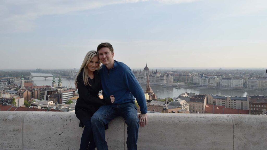 Ashlinn and her husband sitting on a viewpoint overlooking Budapest, the Danube and the houses of parliament