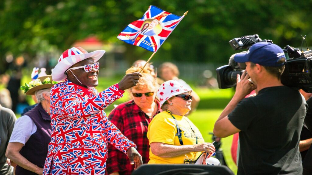 man wearing union jack-printed glasses, hat and jacket, waving a Union Jack flag
