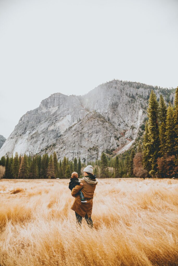 mom and baby standing in a field by some forest and mountain landscapes, taking a break from driving in the family roadtrip