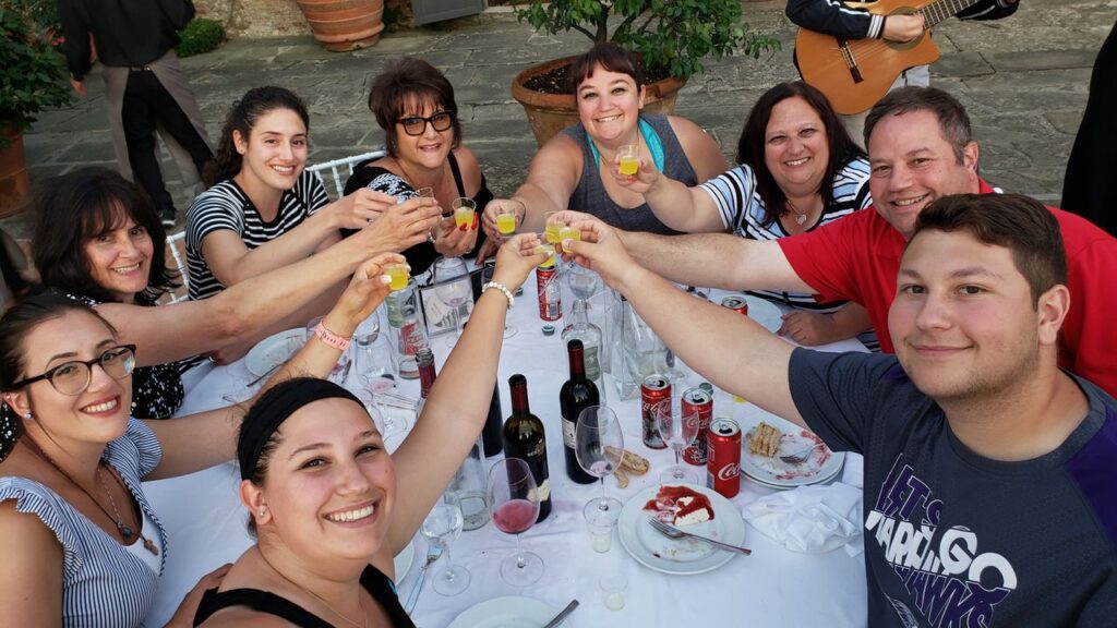A large group of people smile at a dinner table, making a toast with small glasses of post-drink liquer, clinking glasses in the middle of the table.