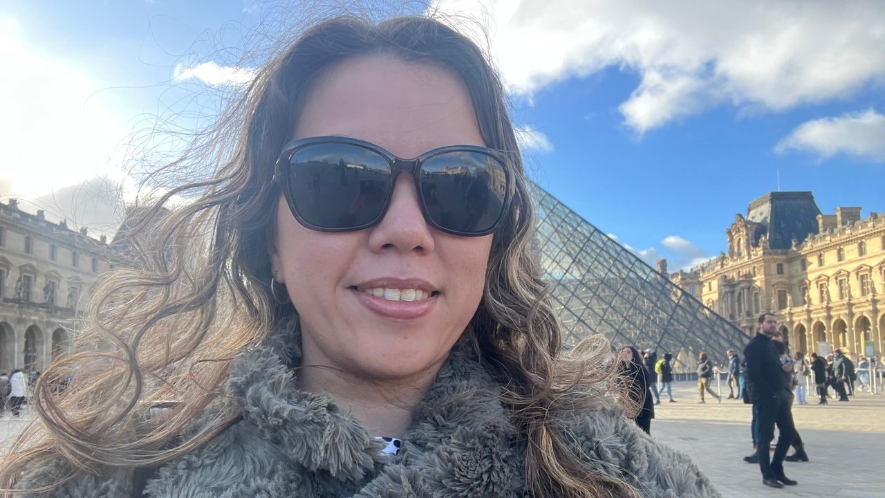 A woman wearing sunglasses smiles for a selfie taken in front of the Louvre