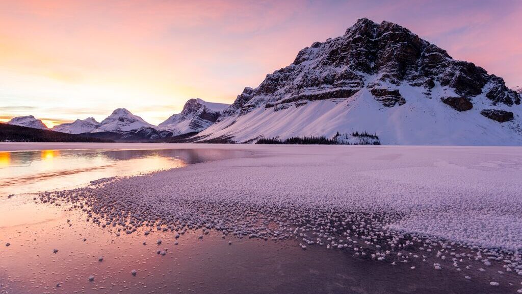 A snowy mountain range in Banff, Canada, at golden hour. The sky is pink and violet, and the ice on the floor reflects these beautiful colors.