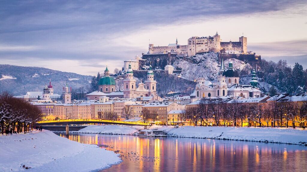 Salzburg during the winter, covered in snow with a slightly frozen river, reflecting the golden lights on the streets. A beautiful place to spend a Christmas holiday