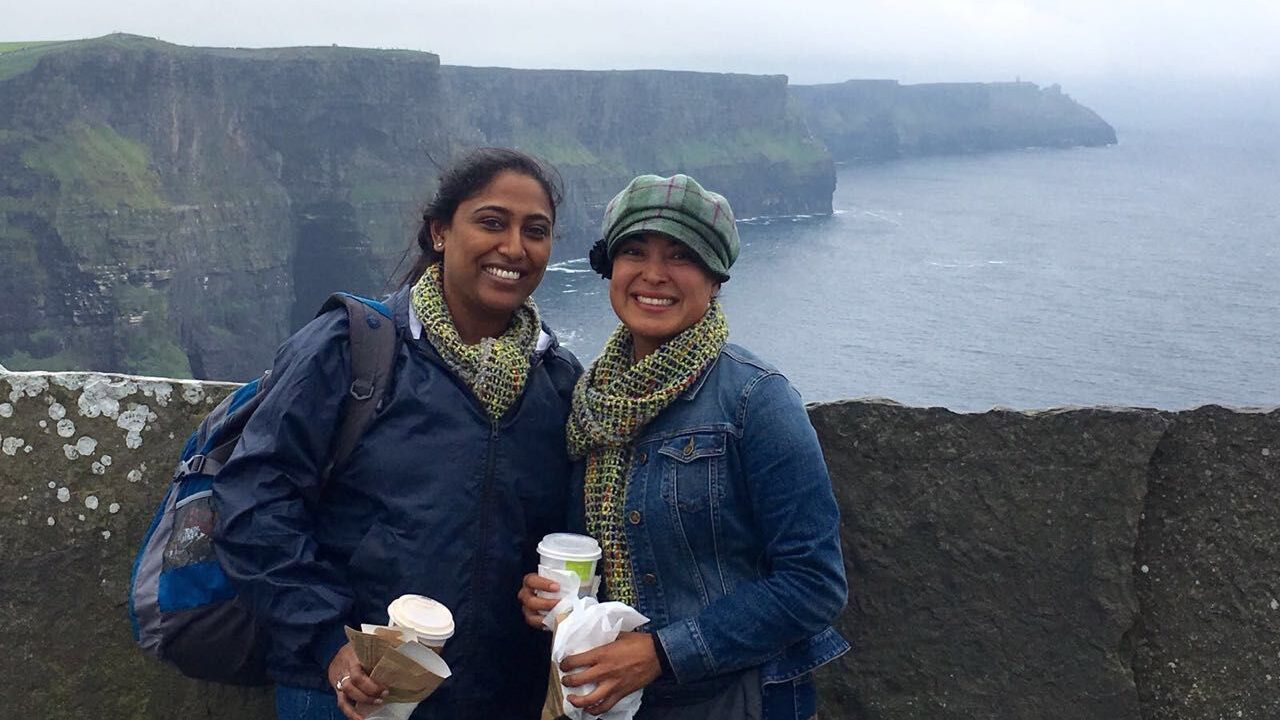 Two friends holding drinks and snacks pose for a photo with vast cliffs and the ocean in the background