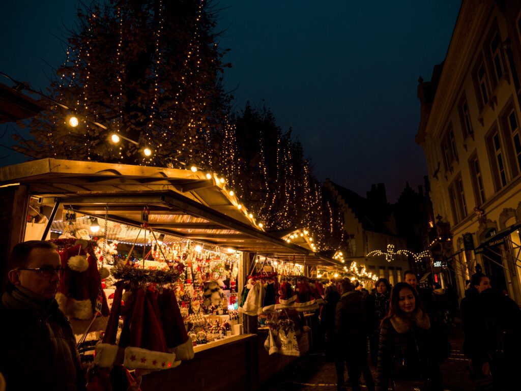 Christmas market stalls lit by twinkling lights at dusk, transformed into a winter wonderland, bustling with shoppers and adorned with festive decorations.