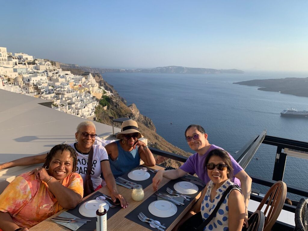 People making friends while traveling by sharing a meal, smiling for the camera with the gorgeous Greek island landscape in the background