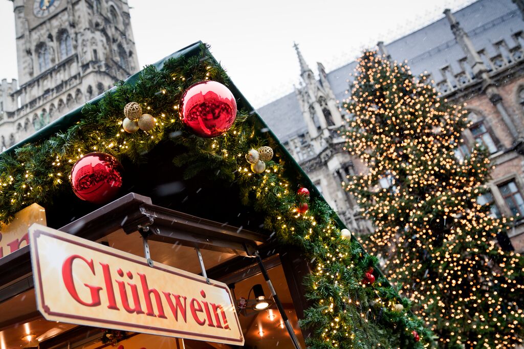 A festive holiday market stall selling glühwein, adorned with large red baubles and garland, with a decorated Christmas tree exuding Christmas charm and a historic building in the background.
