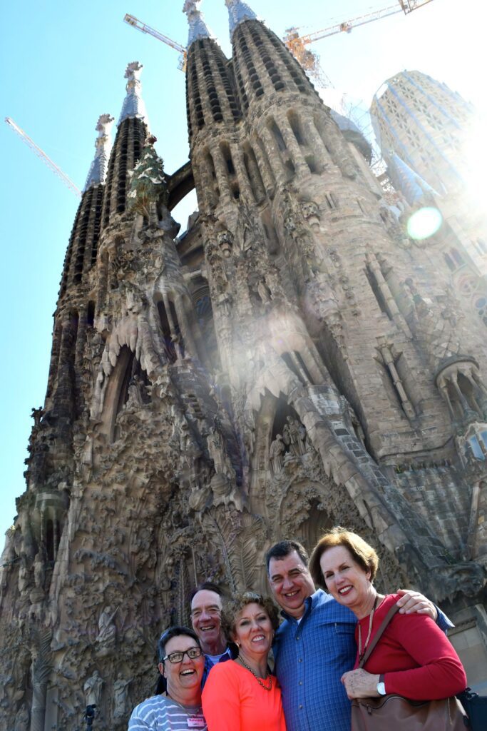 Five adults making friends while traveling, smiling in front of the Sagrada Familia in Barcelona, backlit by the sun with a clear blue sky.